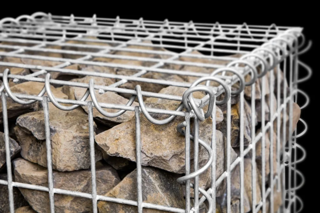 Nahaufnahme einer mit Natursteinen gefüllten Spiralgabione aus verzinktem Stahl mit sichtbaren Spiralverbindern an der Ecke vor schwarzem Hintergrund