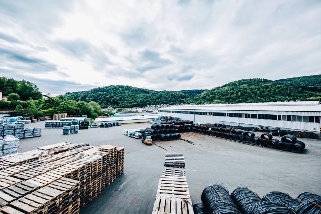 Außengelände eines Industriebetriebs mit gelagerten Stahlcoils, Drahtgeflechtrollen, Paletten und einem Gabelstapler vor bewaldeten Hügeln und Produktionshallen