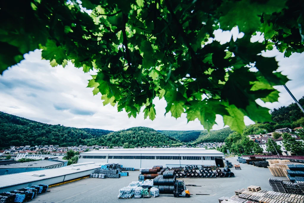 Außengelände eines Industriebetriebs mit gelagerten Stahlcoils und Drahtgeflechtrollen, eingerahmt von Baumästen, mit bewaldeten Hügeln und einer Stadt im Hintergrund